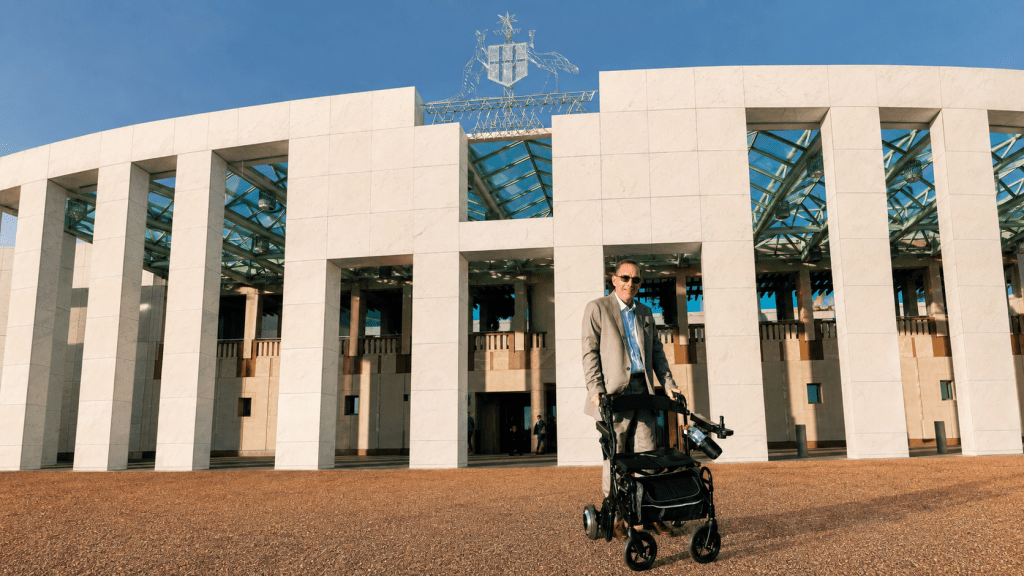 Jeramy Hope standing with his mobility walker in front of Australian Parliament House.